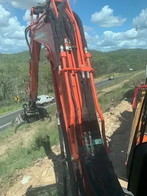 Orange excavator arm working next to a road, with mountains in the background — Boyne Island Tree Services in Boyne Island, QLD
