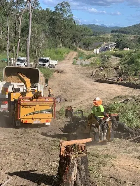 Tree removal: A worker grinds a stump, while a chipper truck processes logs. Rural road setting — Boyne Island Tree Services in Boyne Island, QLD