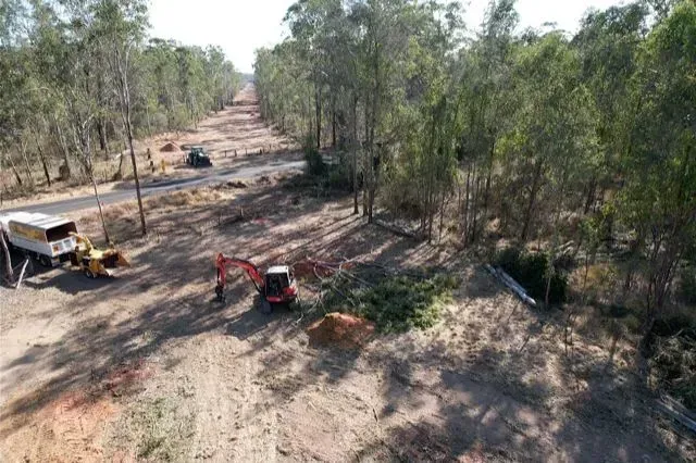 Construction site: Excavator and other equipment clearing trees alongside a dirt road lined with trees — Boyne Island Tree Services in Boyne Island, QLD