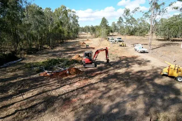 An excavator clearing a dirt path in a wooded area, with service vehicles and a wood chipper nearby — Boyne Island Tree Services in Boyne Island, QLD