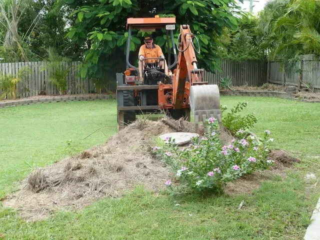 Man operating a backhoe, digging in a grassy yard, orange machinery — Boyne Island Tree Services in Boyne Island, QLD