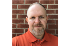 A man in an orange shirt is standing in front of a brick wall