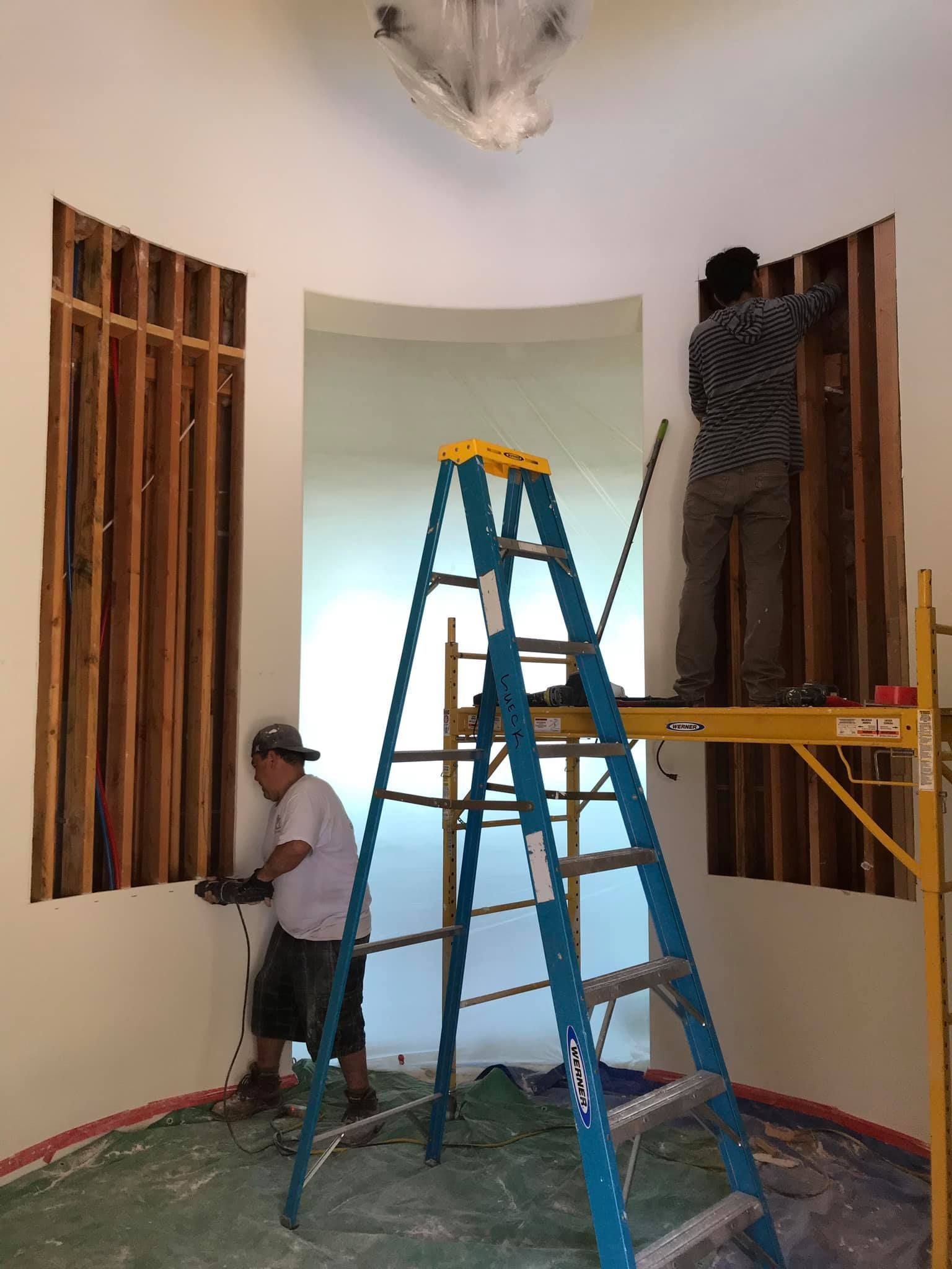Two men are working on a wall in a room with a ladder.