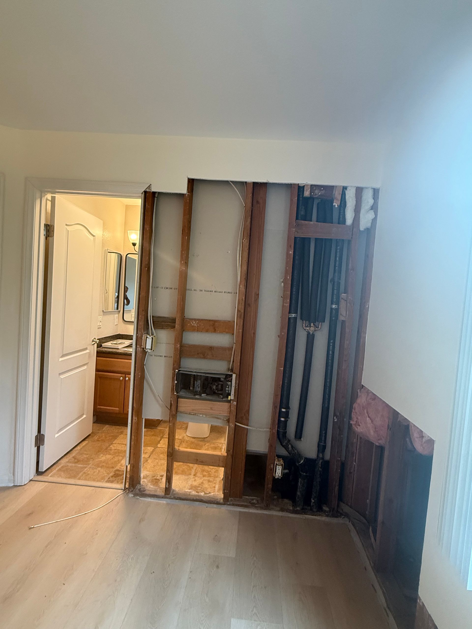 Room with a doorway to a bathroom; a partially demolished wall reveals plumbing and framing. Light-colored wooden flooring.