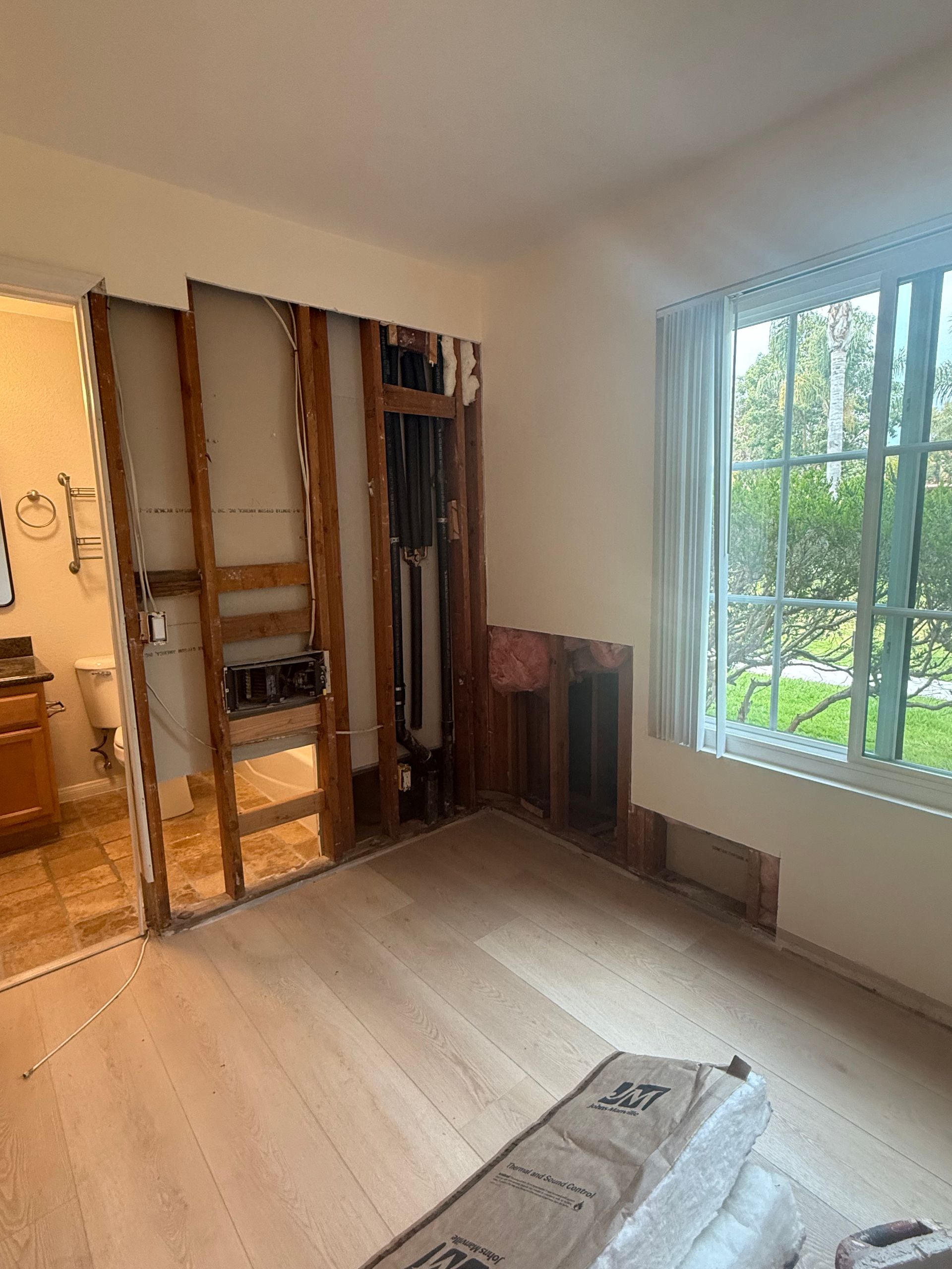 Interior room undergoing renovation with exposed wall studs, new flooring, and a view of a window with trees outside. A bathroom is visible to the left.