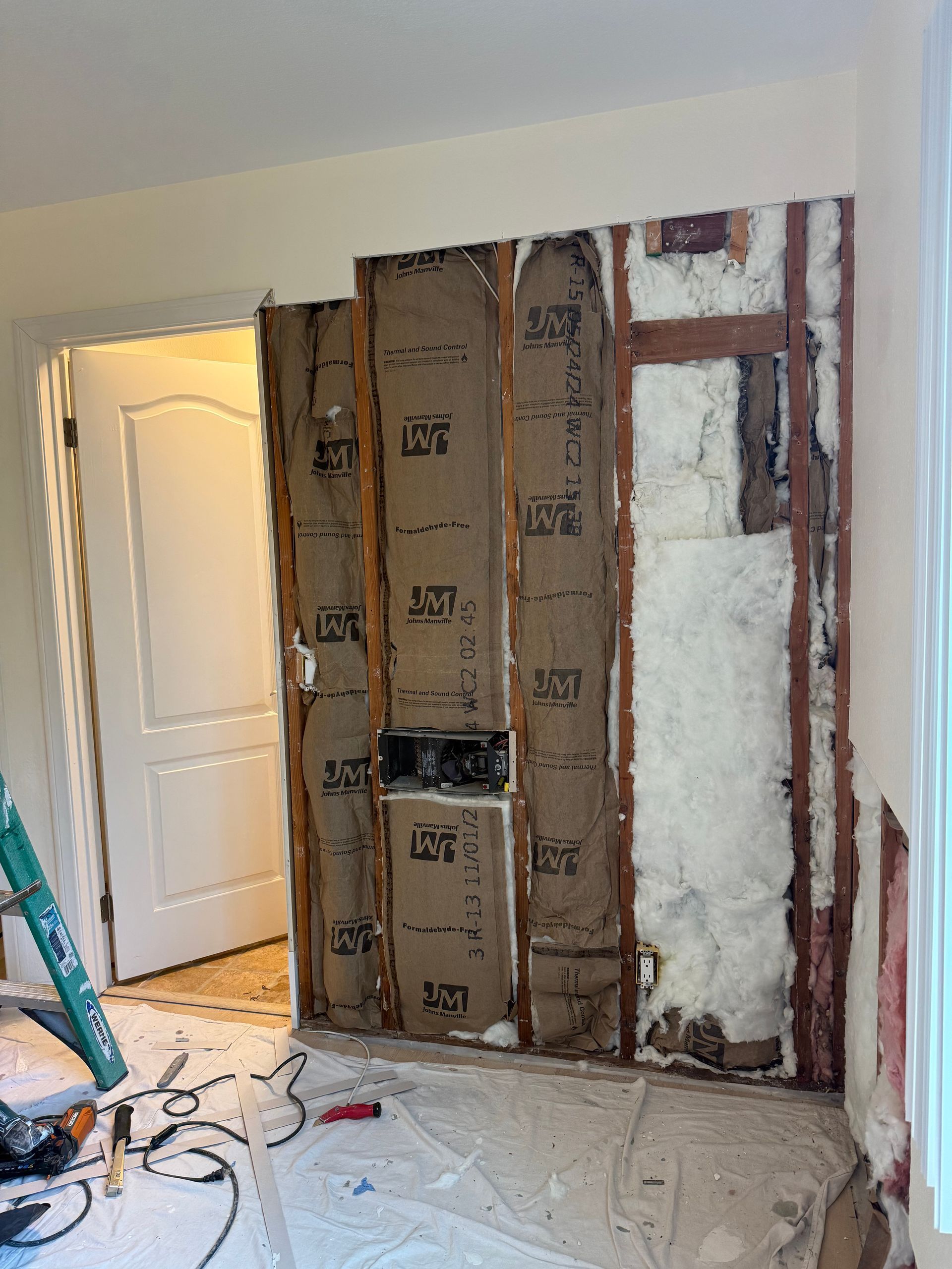 Interior wall under construction with exposed insulation, studs, and electrical components near a doorway.