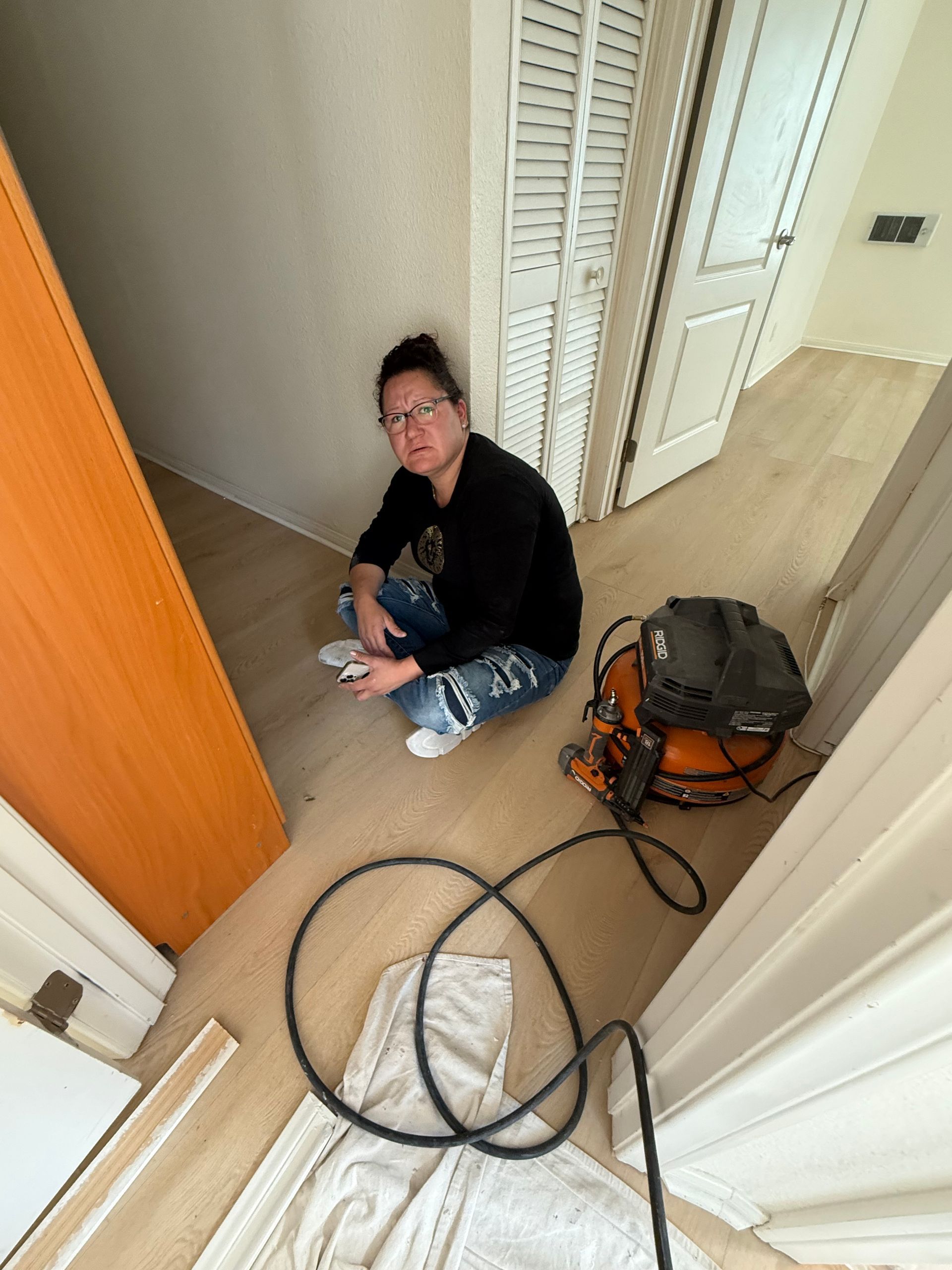 Person sitting on the floor with a vacuum cleaner. They wear a black shirt, jeans, and glasses. The setting is an empty room.