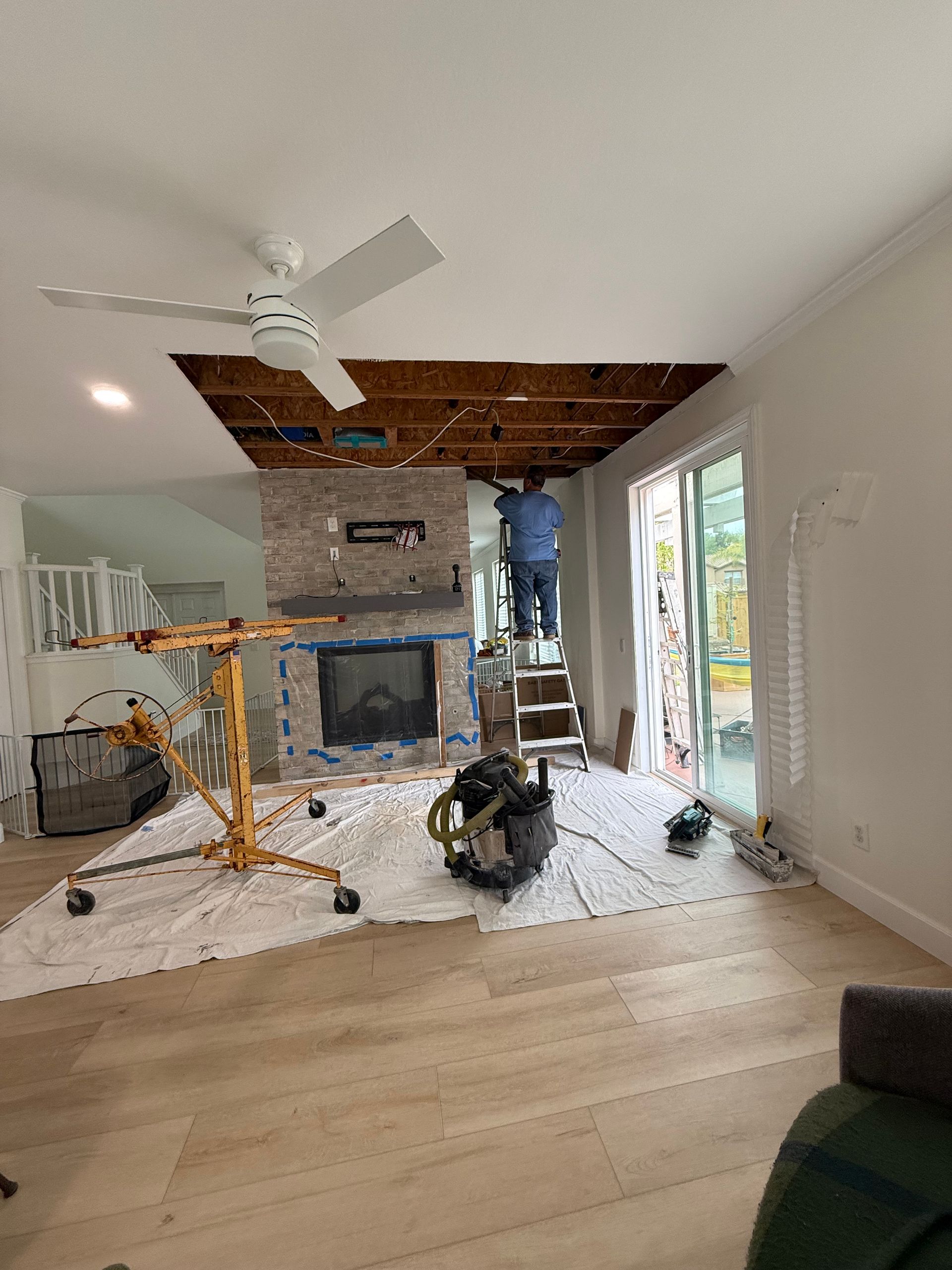 Person on a ladder repairs ceiling above a fireplace. Debris and tools cover the floor.