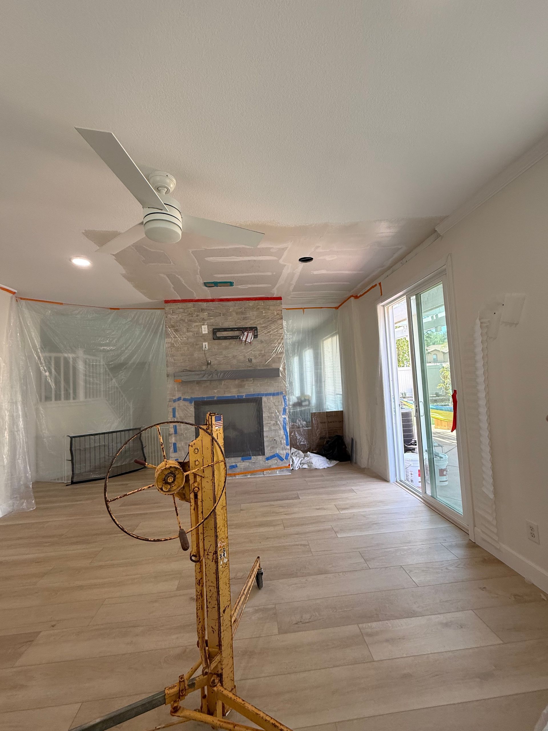 A room under construction with a fireplace, ceiling fan, drywall lift, and light-colored flooring. Protective plastic covers the windows.