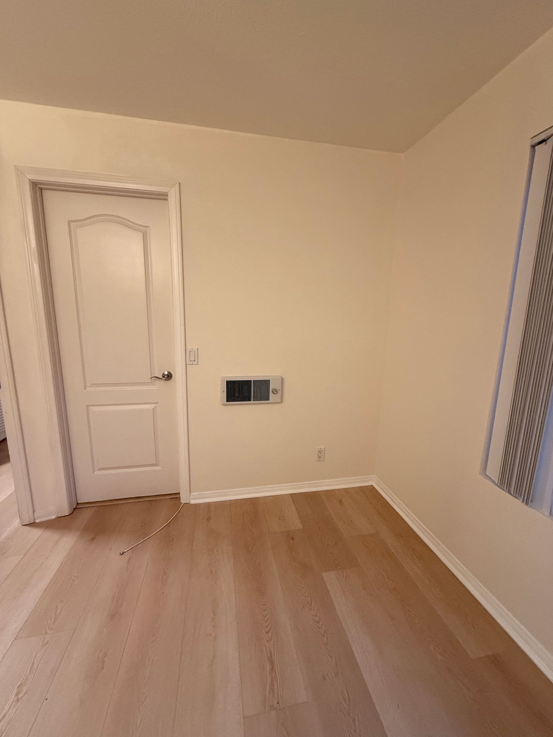 An empty room with light wood flooring, beige walls, a closed white door, and a window with a curtain.