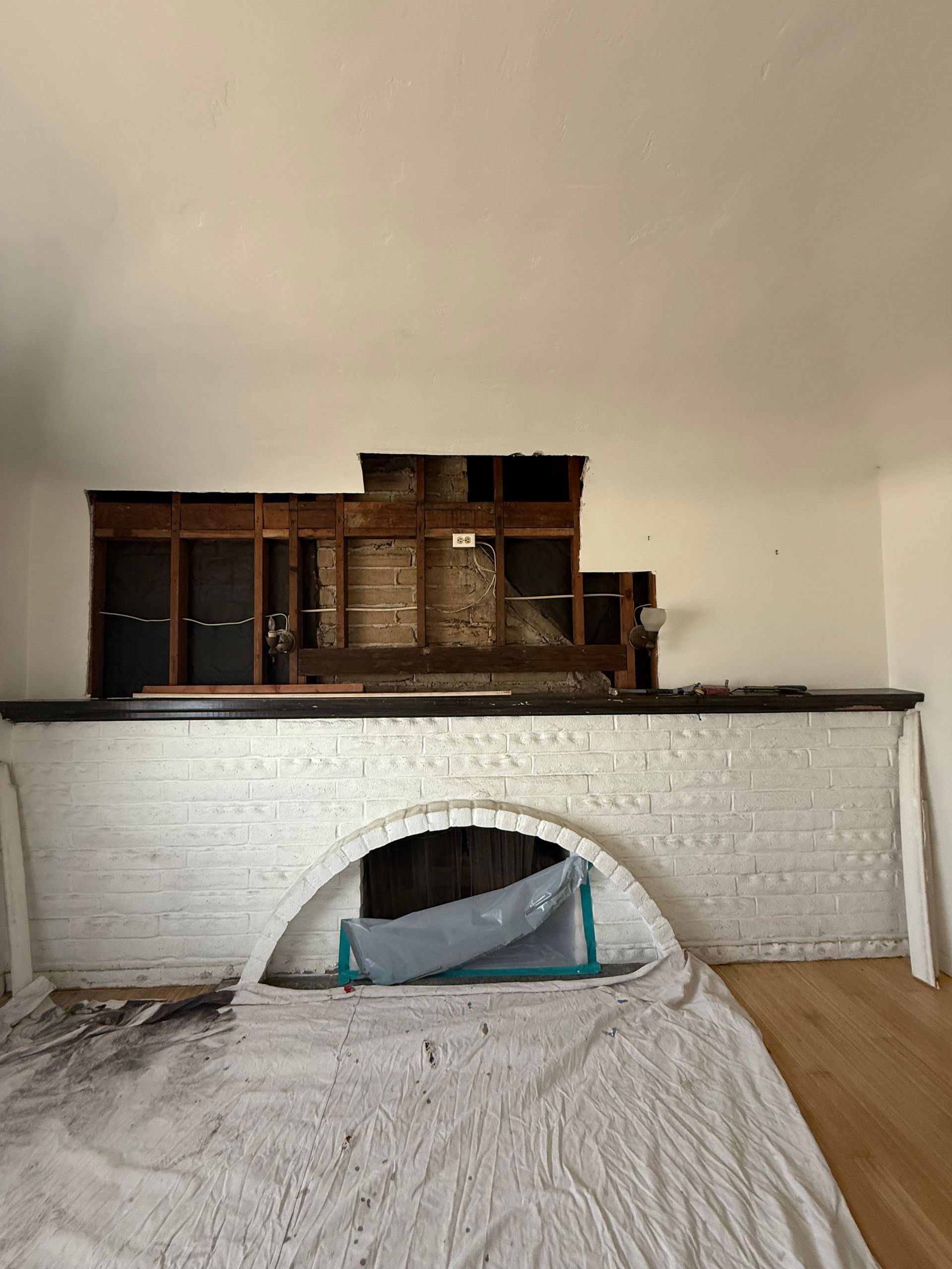 Fireplace with white brick surround, an arched opening, and exposed wooden structure above. A white drop cloth covers the floor in front.