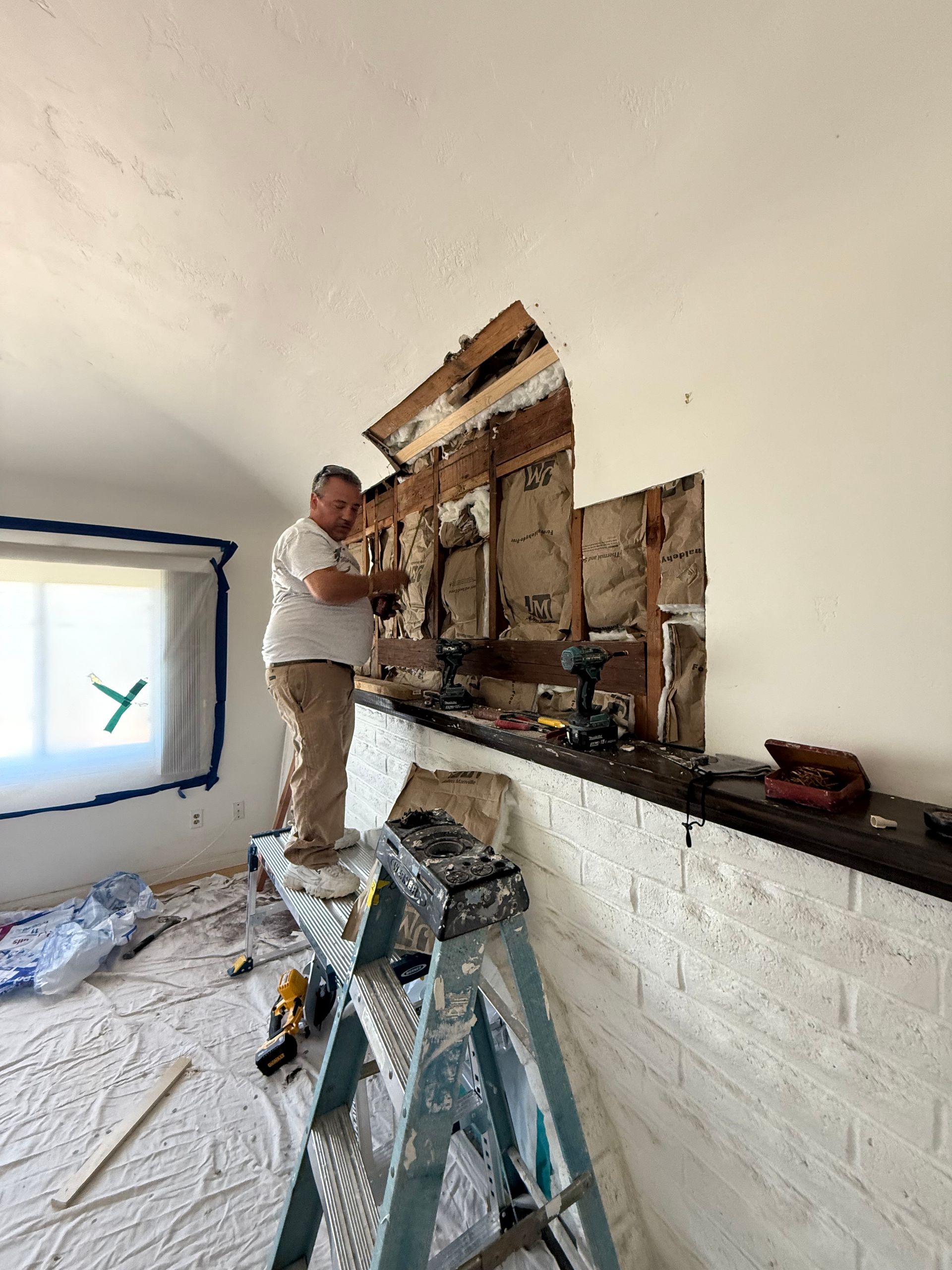 A man stands on a ladder, demolishing a wall section in a room with white-painted brick. He wears work clothes. The room is under renovation.