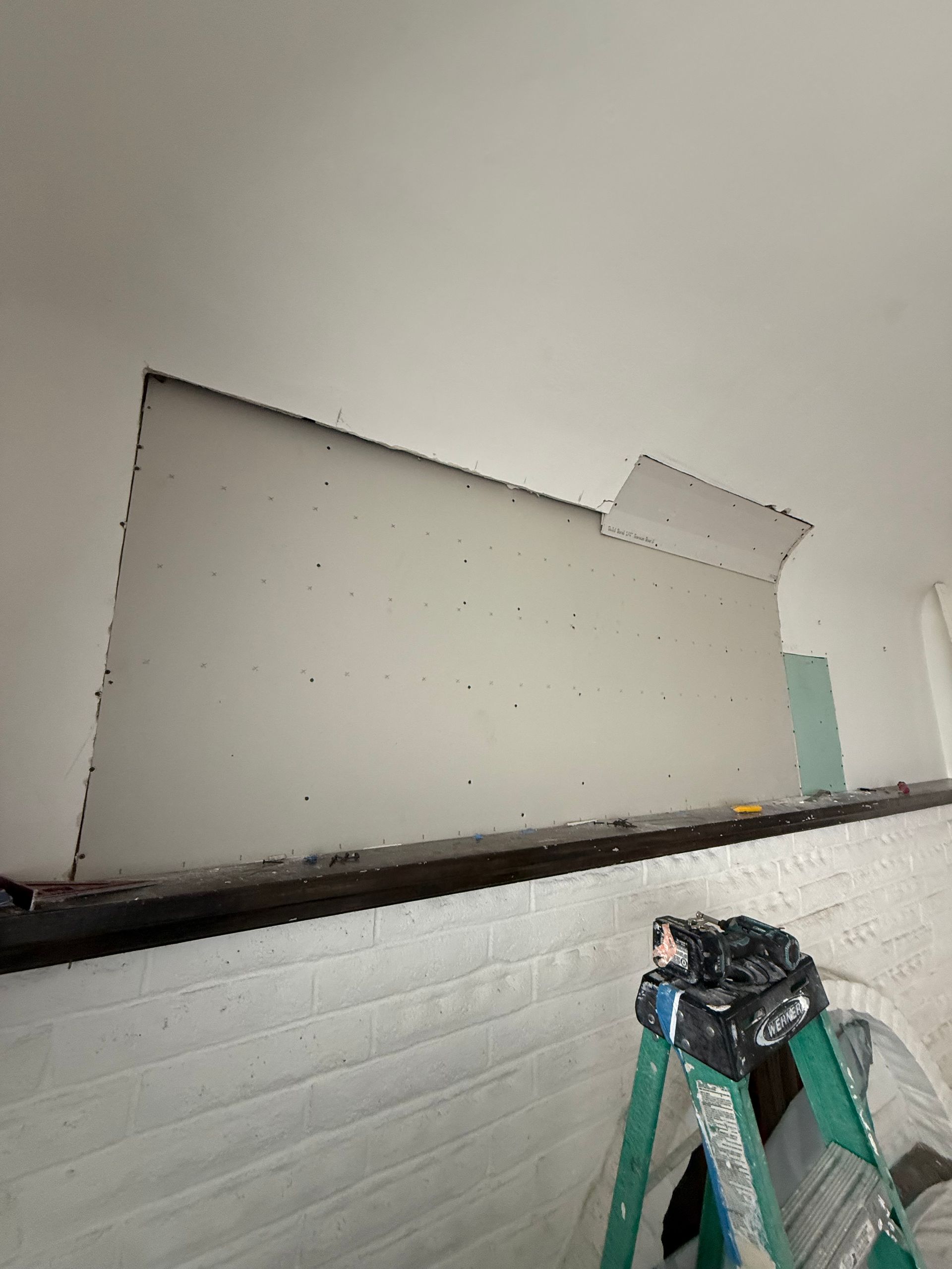 Drywall repair in progress; section of ceiling and wall with cut drywall, ladder, and brick wall below.