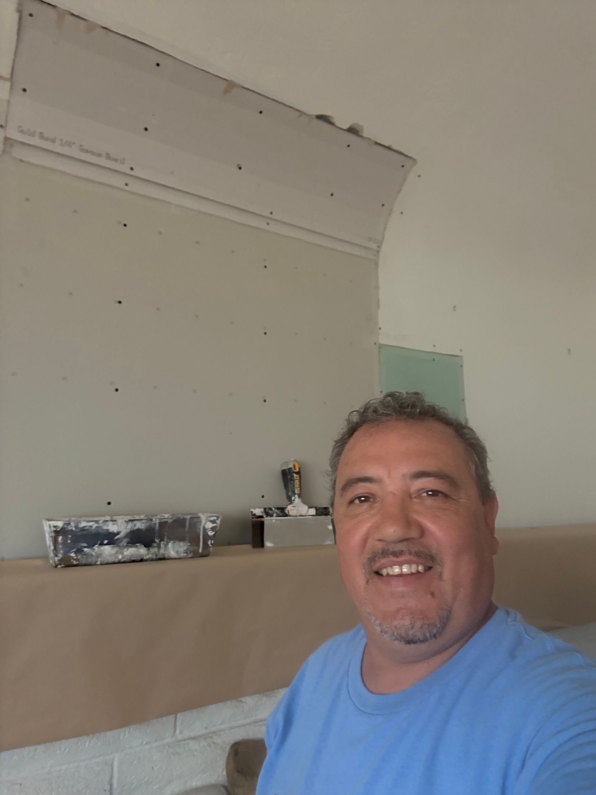 A smiling man stands in a room with drywall construction. He's wearing a blue shirt. Beige paper protects a wall.