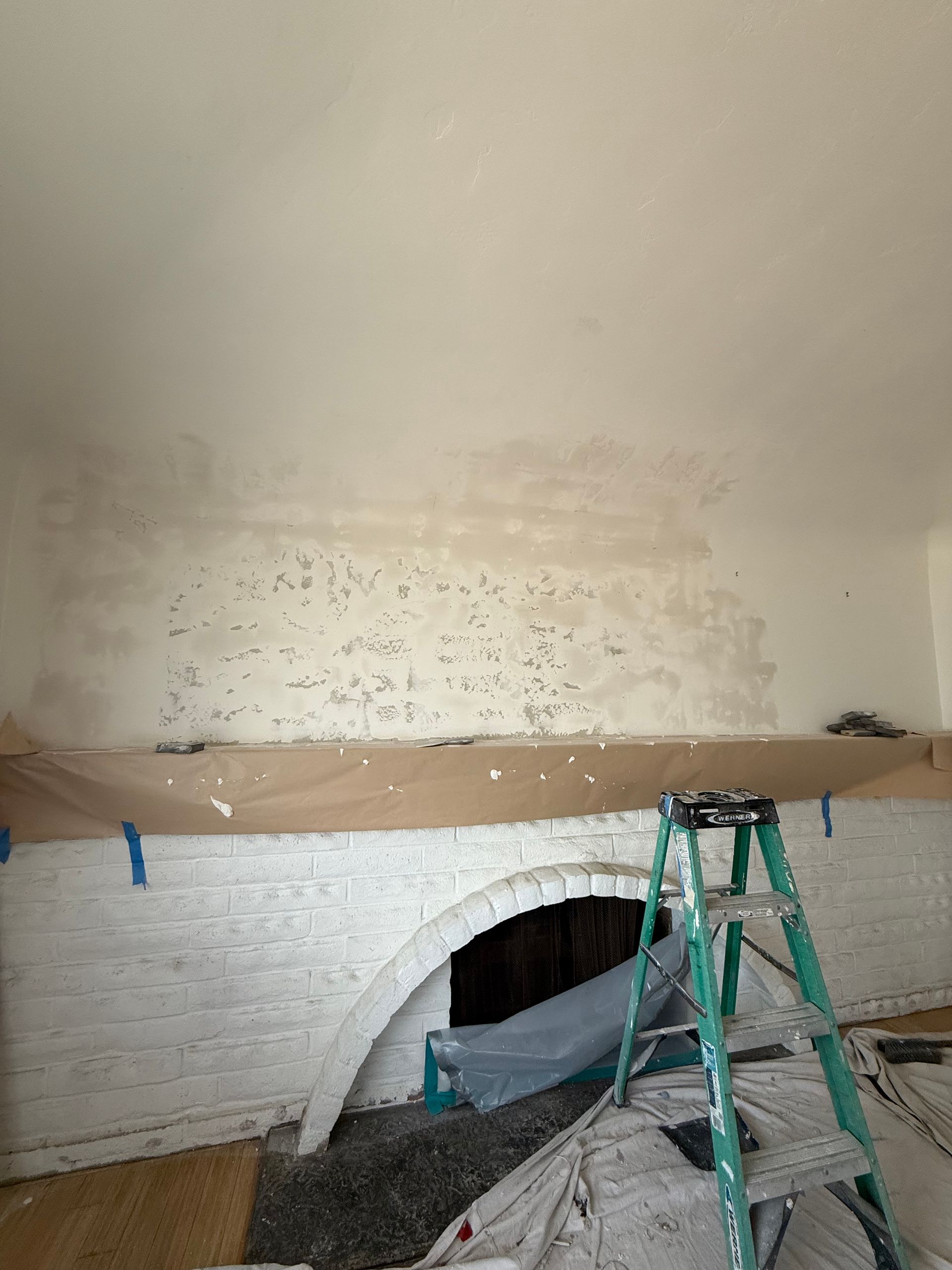 A white painted brick fireplace with an arched opening is being prepared for painting. A ladder is set up near it.