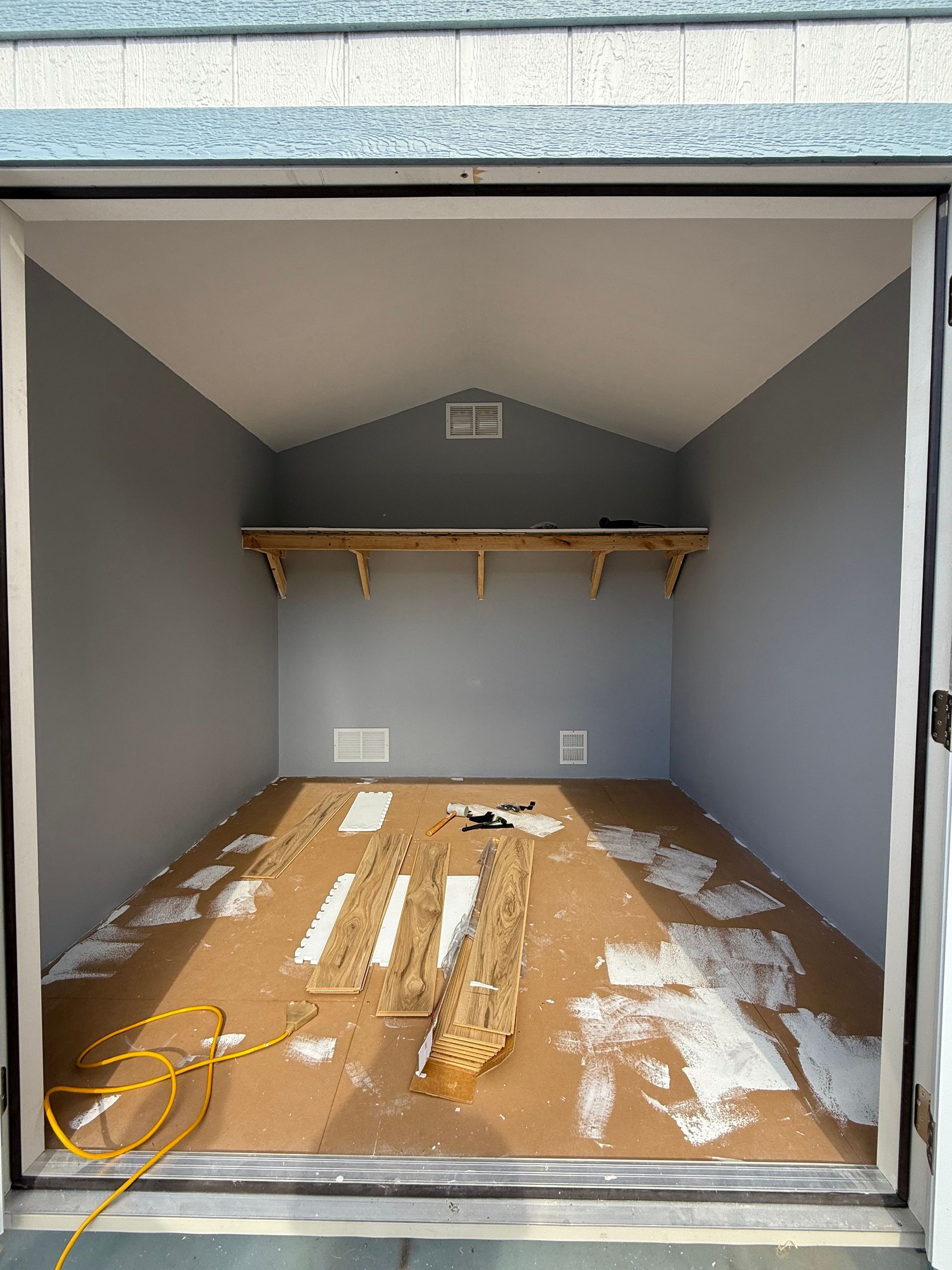 Inside of an empty shed with gray walls and a wooden floor. A shelf is mounted on the back wall.