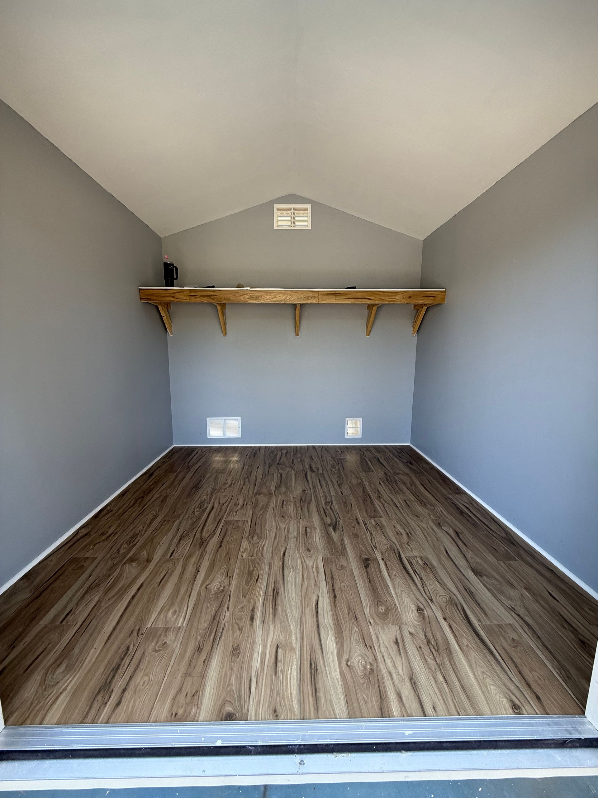 Interior of a small room with wood-look flooring, gray walls, a shelf, and a vent.