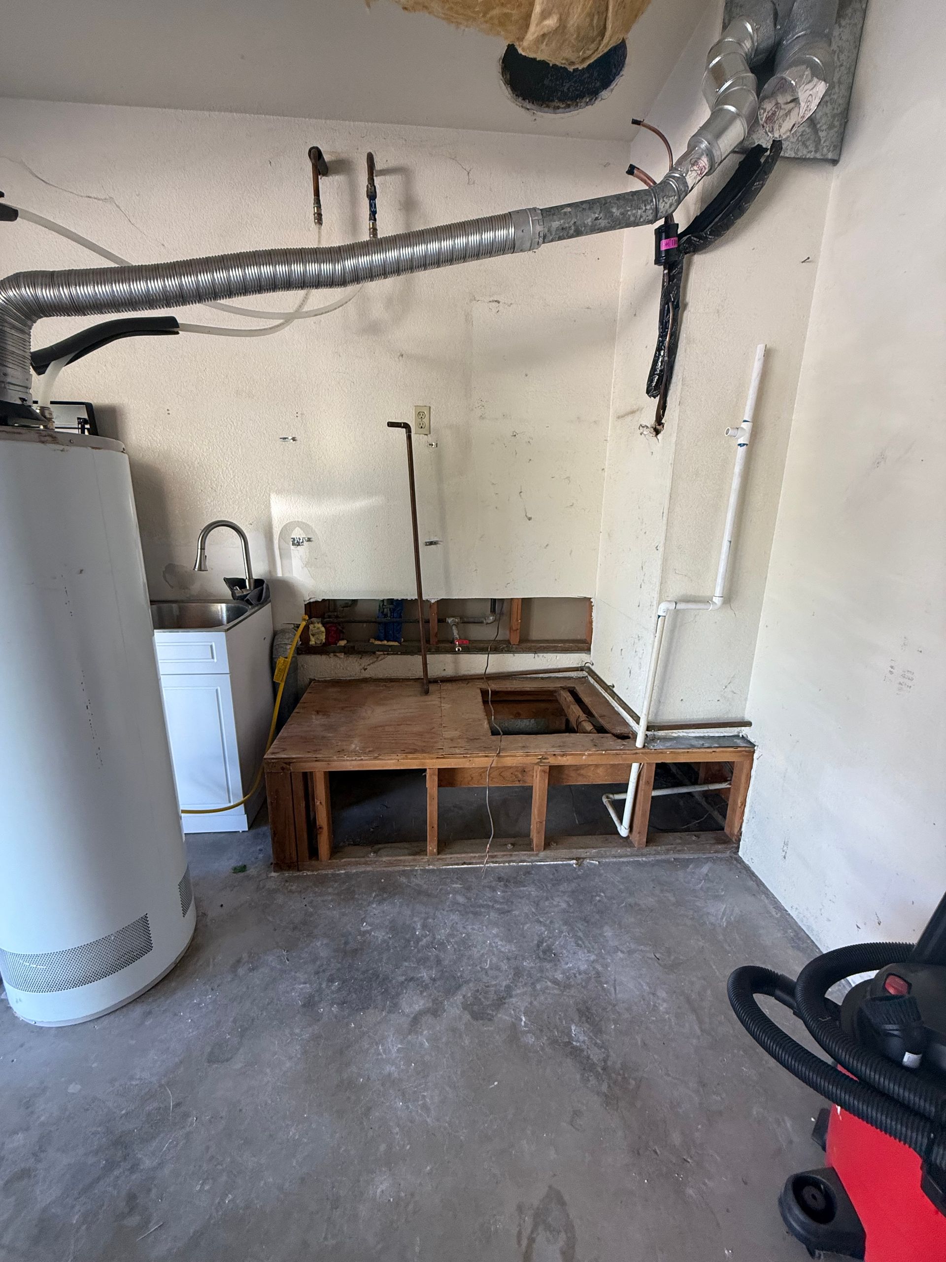 Laundry room with exposed wooden frame, water heater, sink, ductwork, and vacuum. Interior shot.