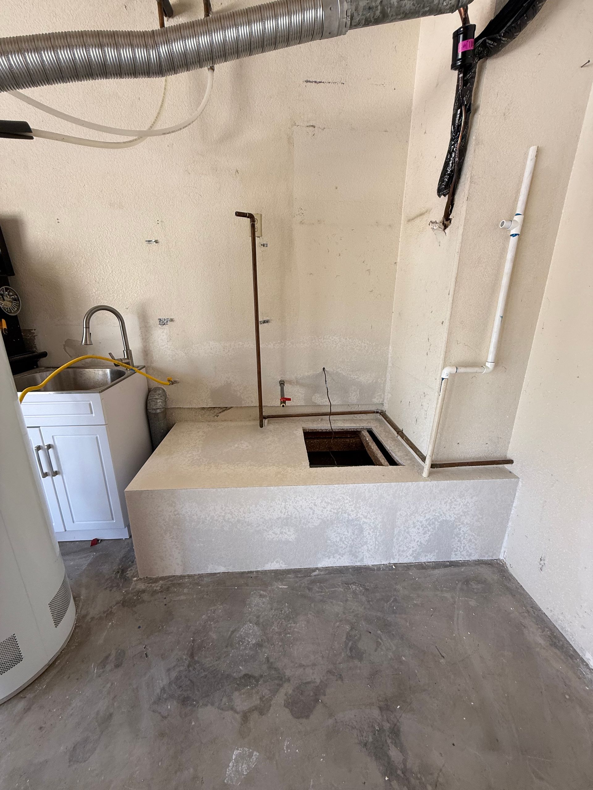 A concrete structure with an open square hole, plumbing pipes, and a small sink. Located indoors, likely a utility room.