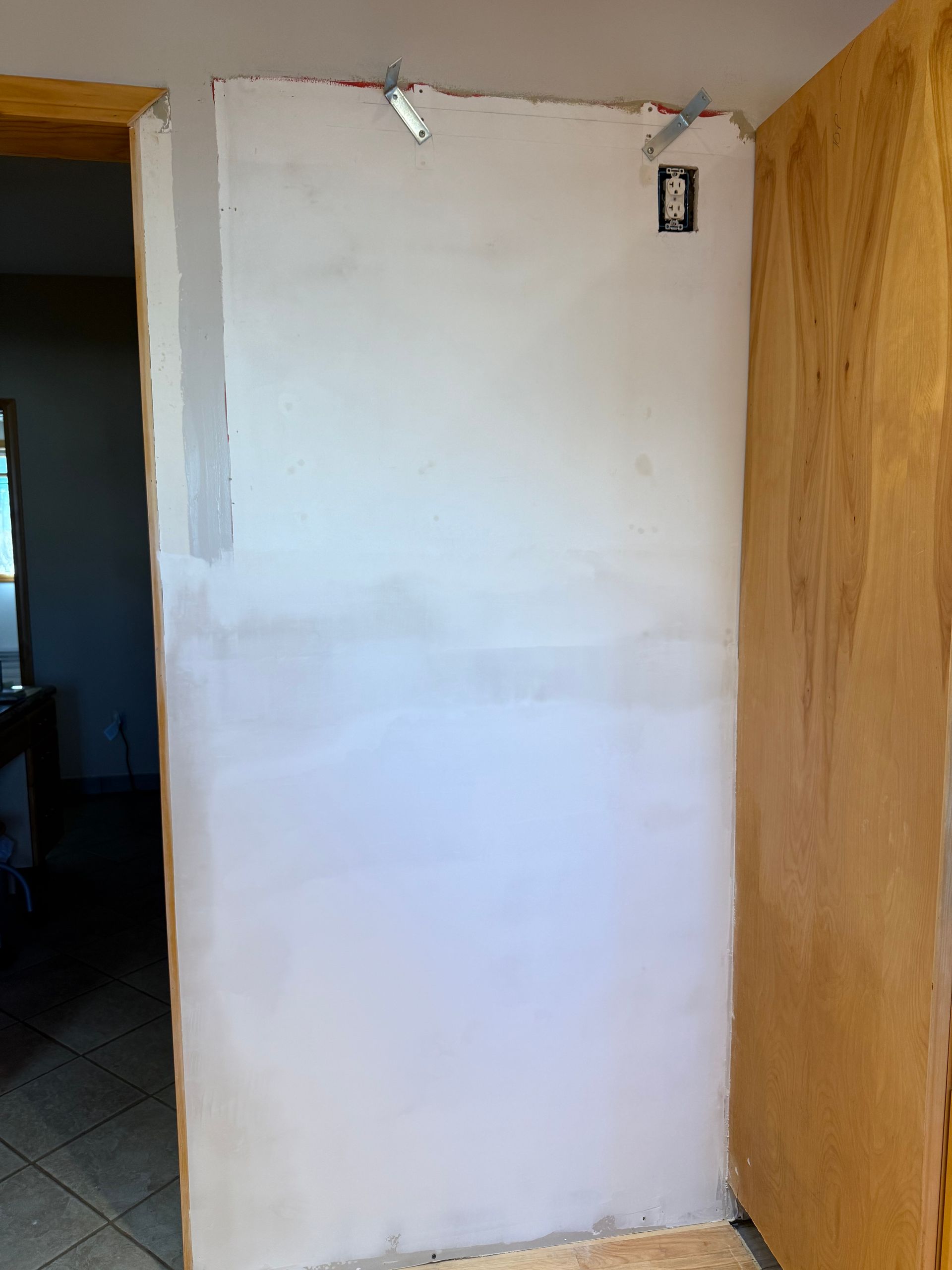 White drywall on a wall with a doorway on the left and a wooden cabinet on the right, an electrical outlet, and construction clips.
