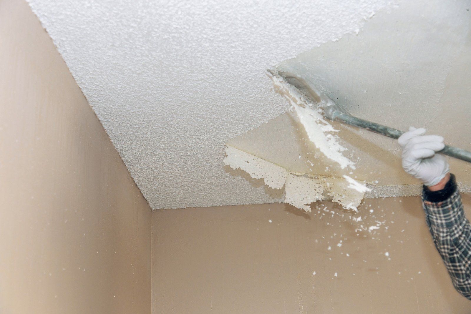 Person scrapes textured ceiling with a scraper, removing popcorn texture. Beige walls surround.