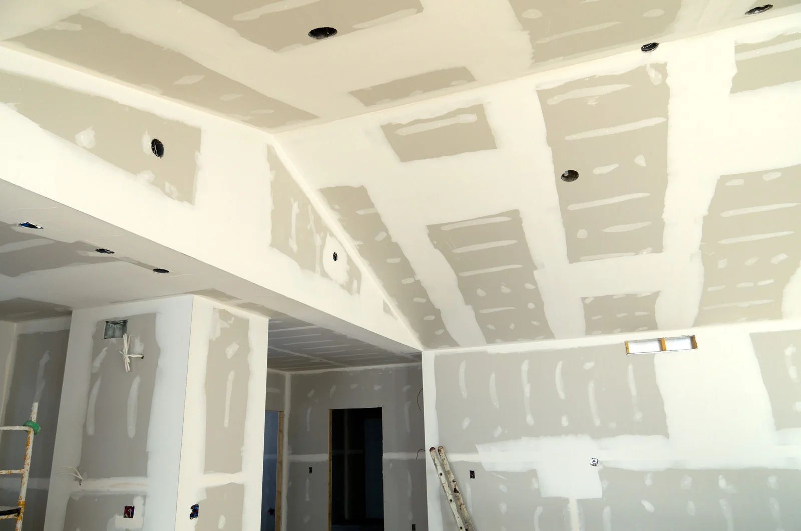 Interior of a room under construction, with drywall installed on walls and ceiling; recessed lighting fixtures.