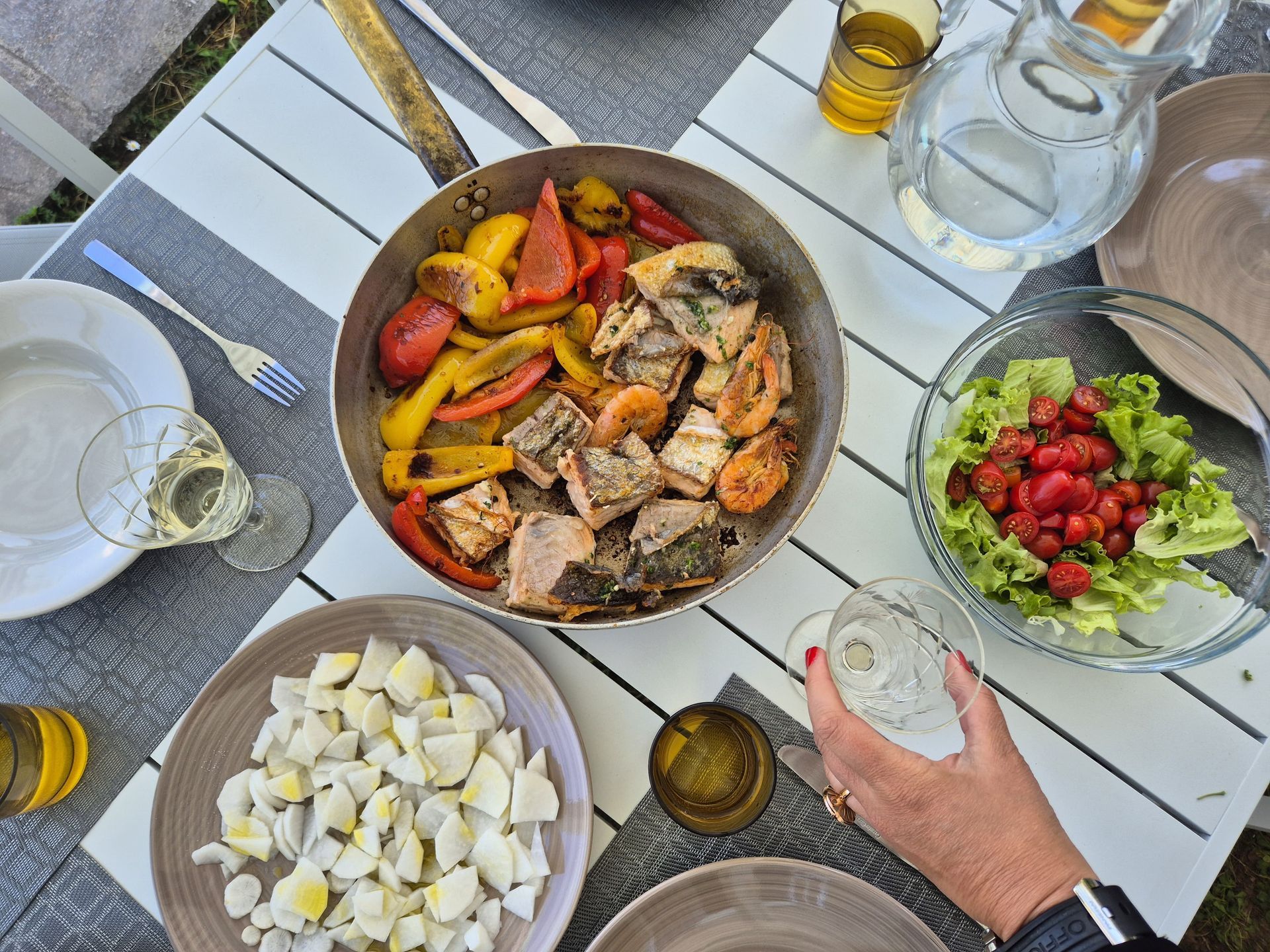 Draufsicht auf eine Mahlzeit: Pfanne mit Essen, Salat, Eierscheiben, Gläser und eine Hand, die nach einem Glas greift.
