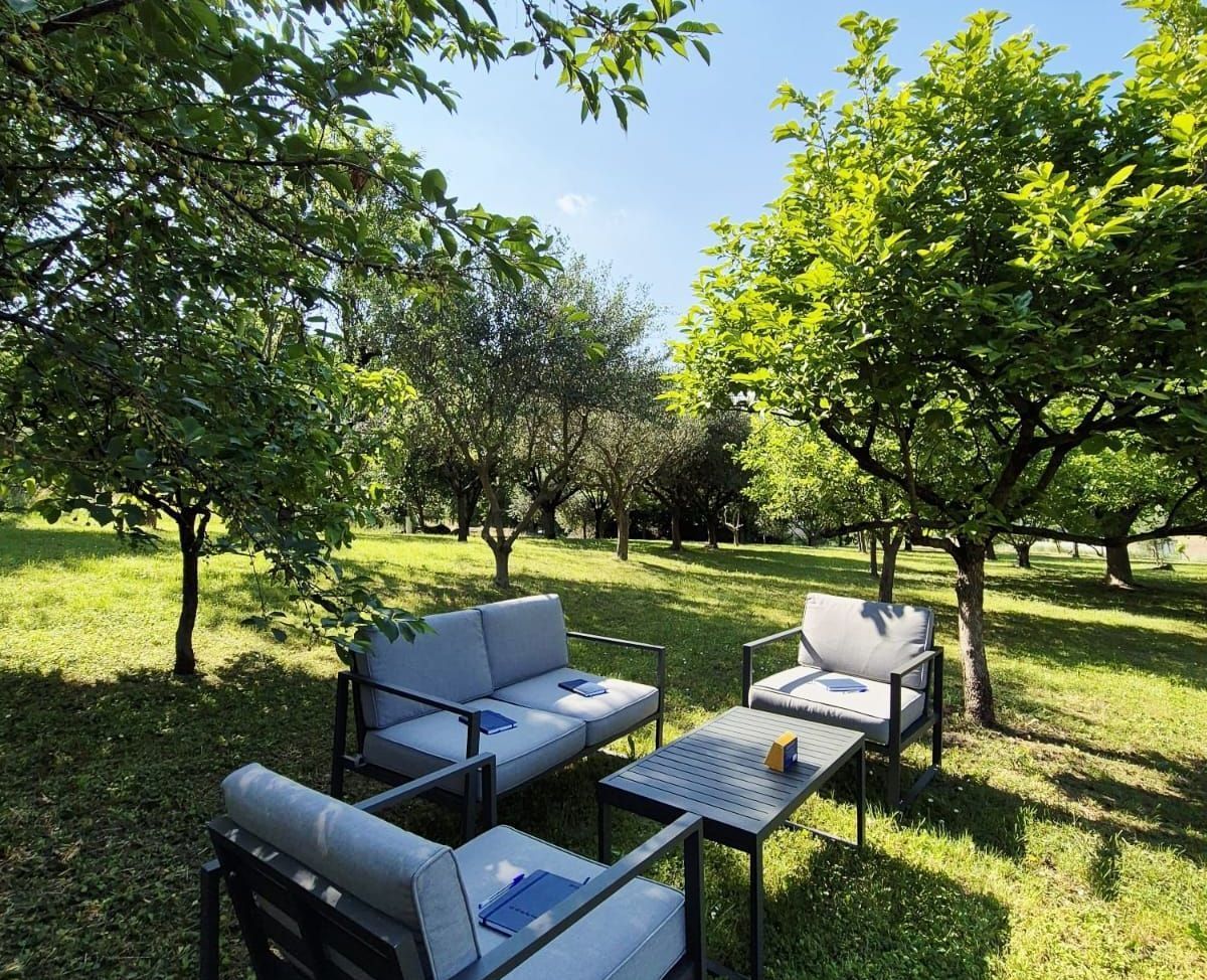 Sitzbereich im Freien mit grauen Möbeln unter grünen Bäumen an einem sonnigen Tag.