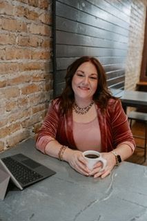 Woman in pink blazer smiles, holding coffee at a cafe table with a laptop, brick wall background.