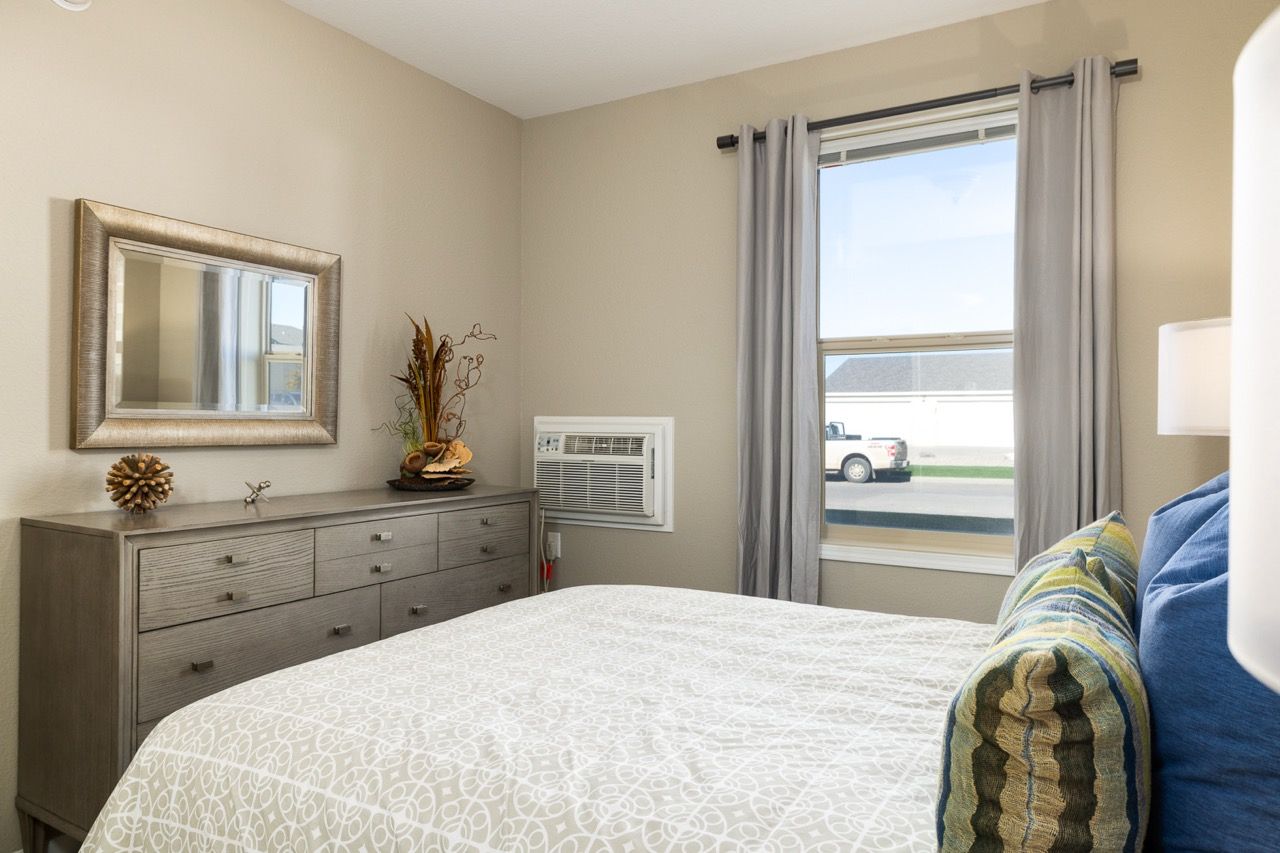 Bedroom in apartment with bed, dresser, mirror, window and curtains; wall AC unit.