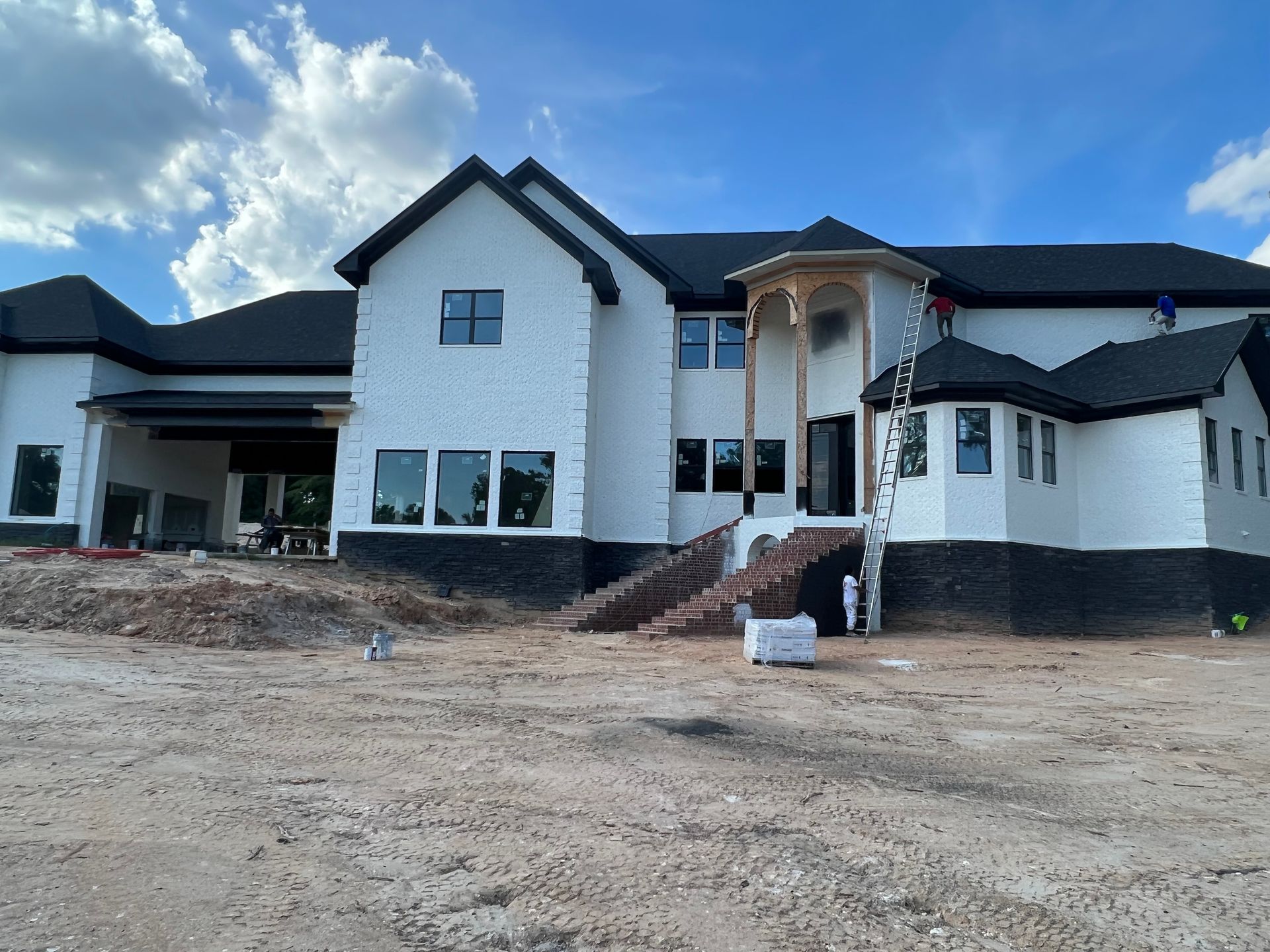 Large white brick house under construction, black roof, steps, and exposed dirt.