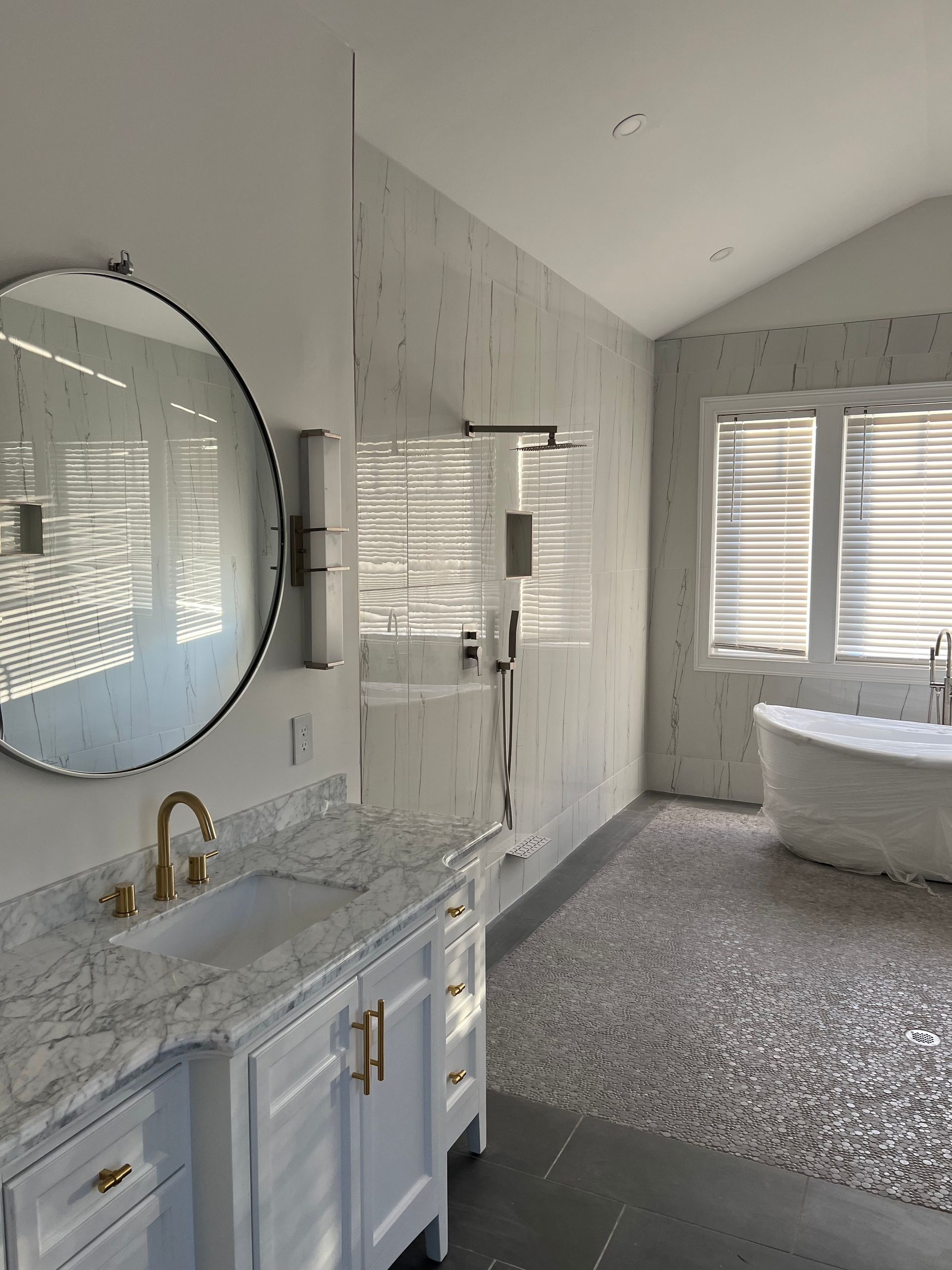 Bathroom with vanity, large mirror, shower, and freestanding tub. Gray pebble floor. White walls with windows.
