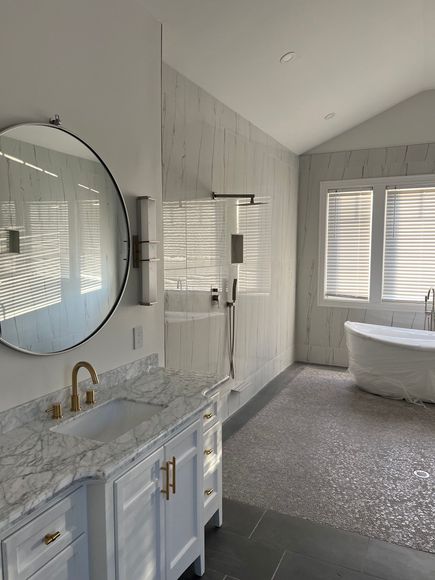 Bathroom with vanity, large mirror, shower, and freestanding tub. Gray pebble floor. White walls with windows.