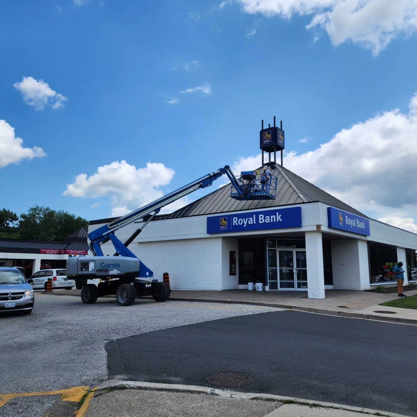 A construction lift reaches toward the roof of a Royal Bank building under a blue sky with white clouds.