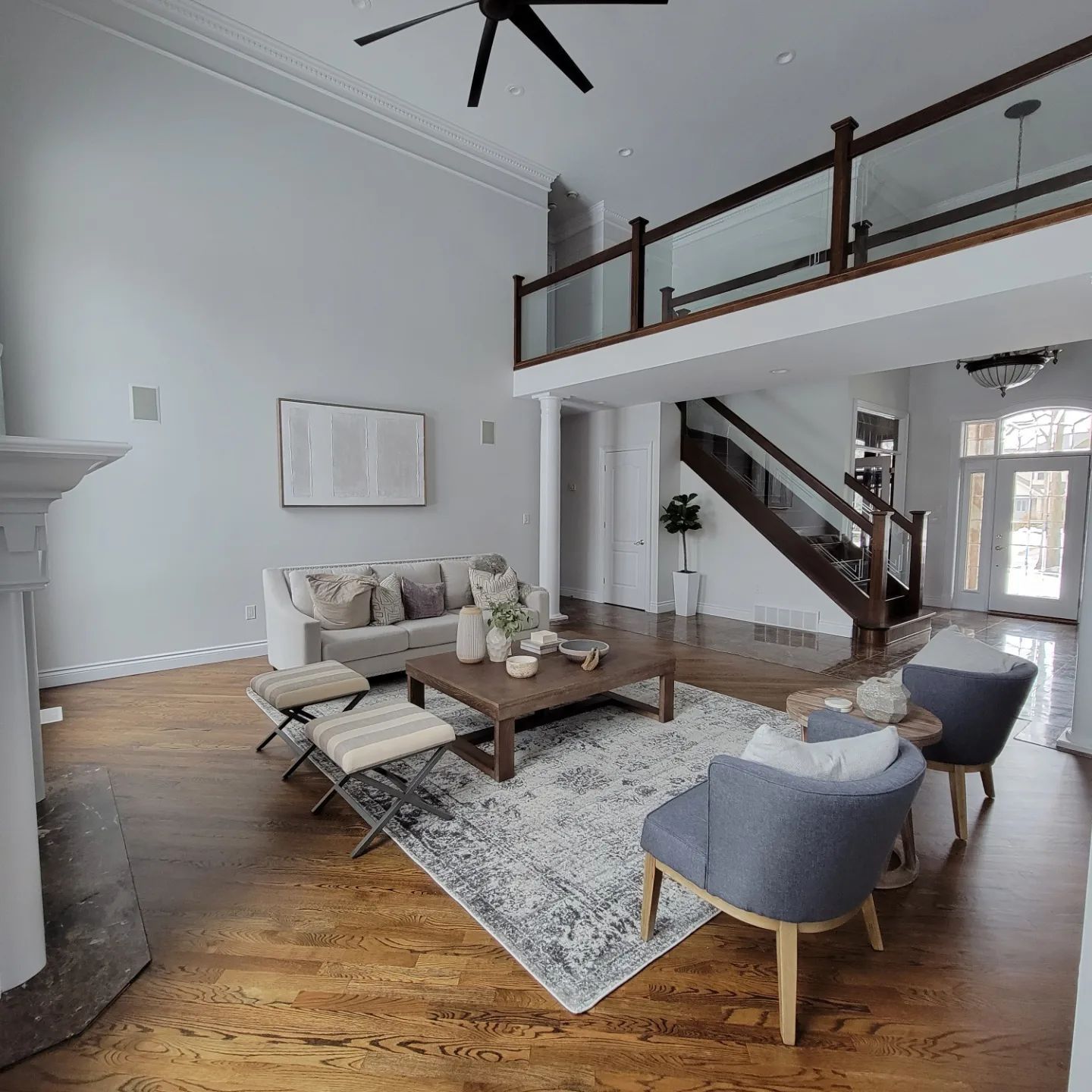 Modern, high-ceiling living room with wood floors, a neutral sofa, patterned rug, armchairs, and a wooden staircase.