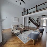 Modern, high-ceiling living room with wood floors, a neutral sofa, patterned rug, armchairs, and a wooden staircase.