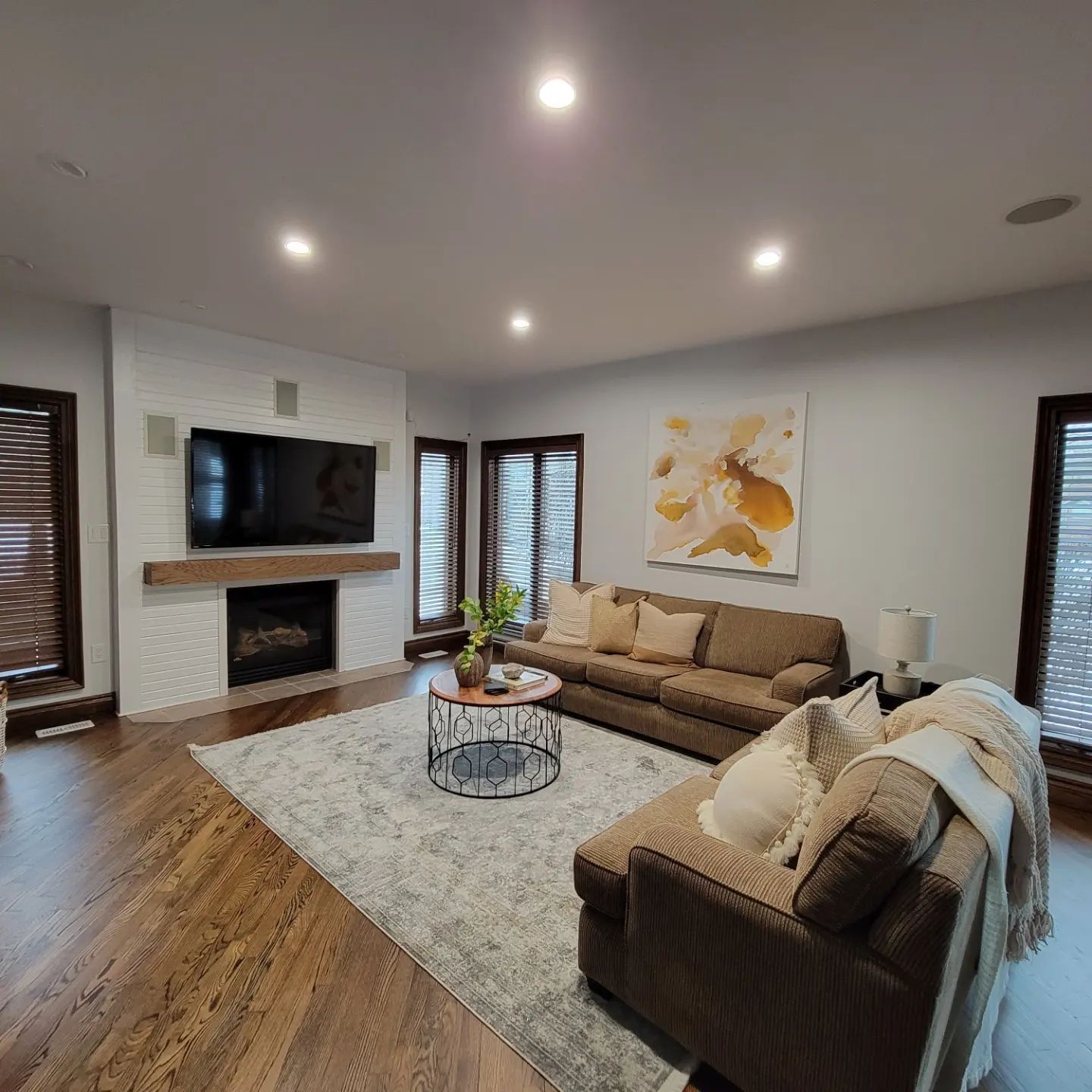 A living room with a brick fireplace, television, brown sectional sofa, area rug, and abstract art on a light wall.