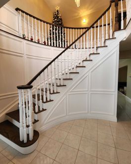 A curved staircase with dark wooden steps and white railings features ornate paneling and a decorated Christmas tree.