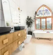 A luxury bathroom with a fluted wooden vanity, black sinks, a freestanding clawfoot tub, and an arched stained-glass window.