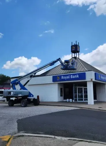 A construction lift reaches toward the roof of a Royal Bank building under a blue sky with white clouds.