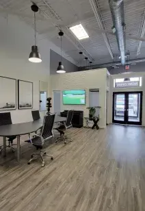 A modern conference room with a gray oval table, four black chairs, hanging lights, a wall-mounted TV, and glass doors.