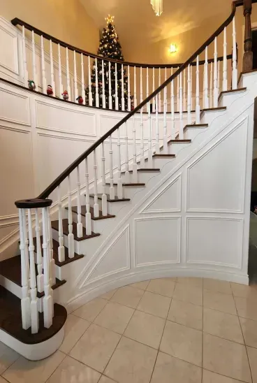 A curved staircase with dark wooden steps and white railings features ornate paneling and a decorated Christmas tree.