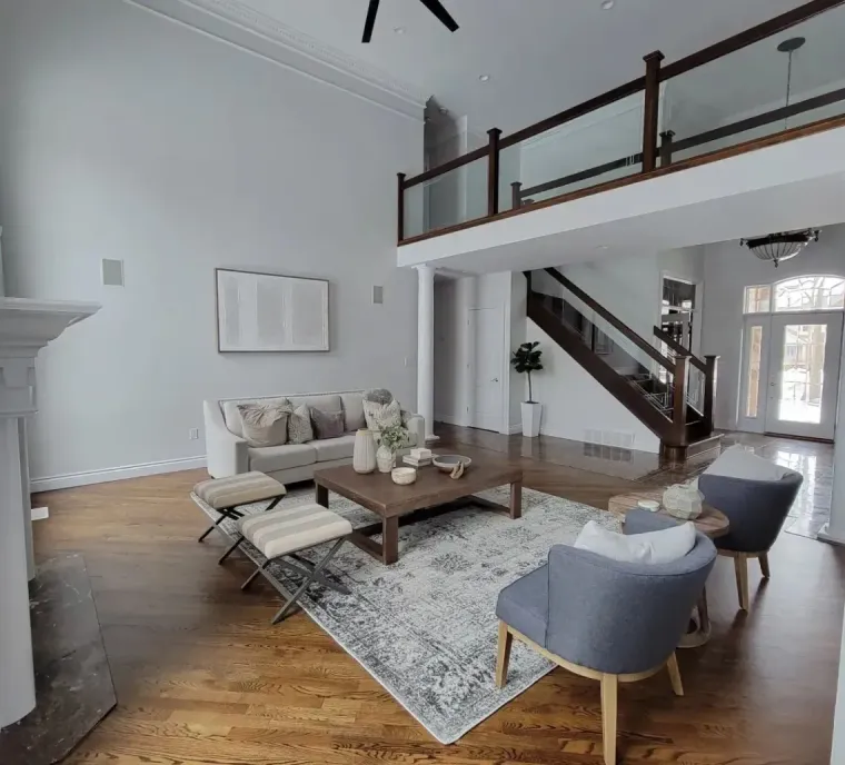 A spacious, modern living room with hardwood floors, a gray sofa, coffee table, blue armchairs, and a lofted balcony.