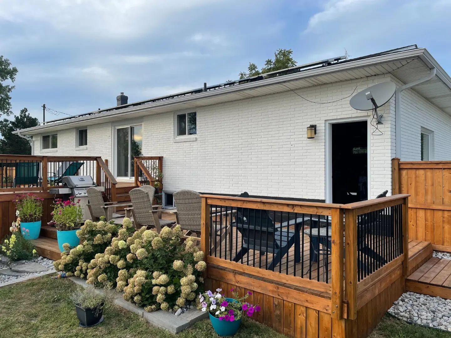 A white brick house with a large wooden deck, patio furniture, and white hydrangea bushes in the backyard.