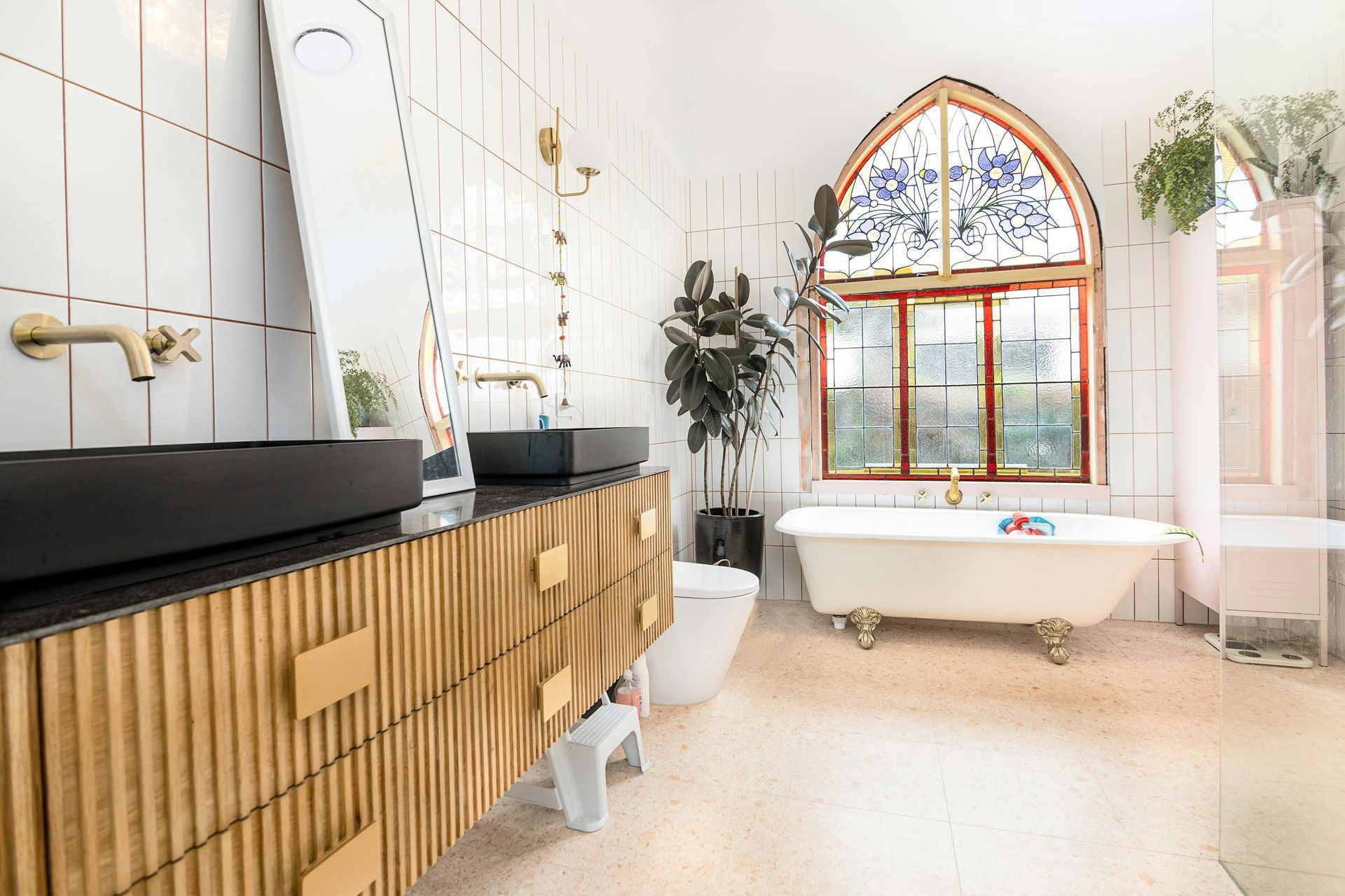 A luxury bathroom with a fluted wooden vanity, black sinks, a freestanding clawfoot tub, and an arched stained-glass window.