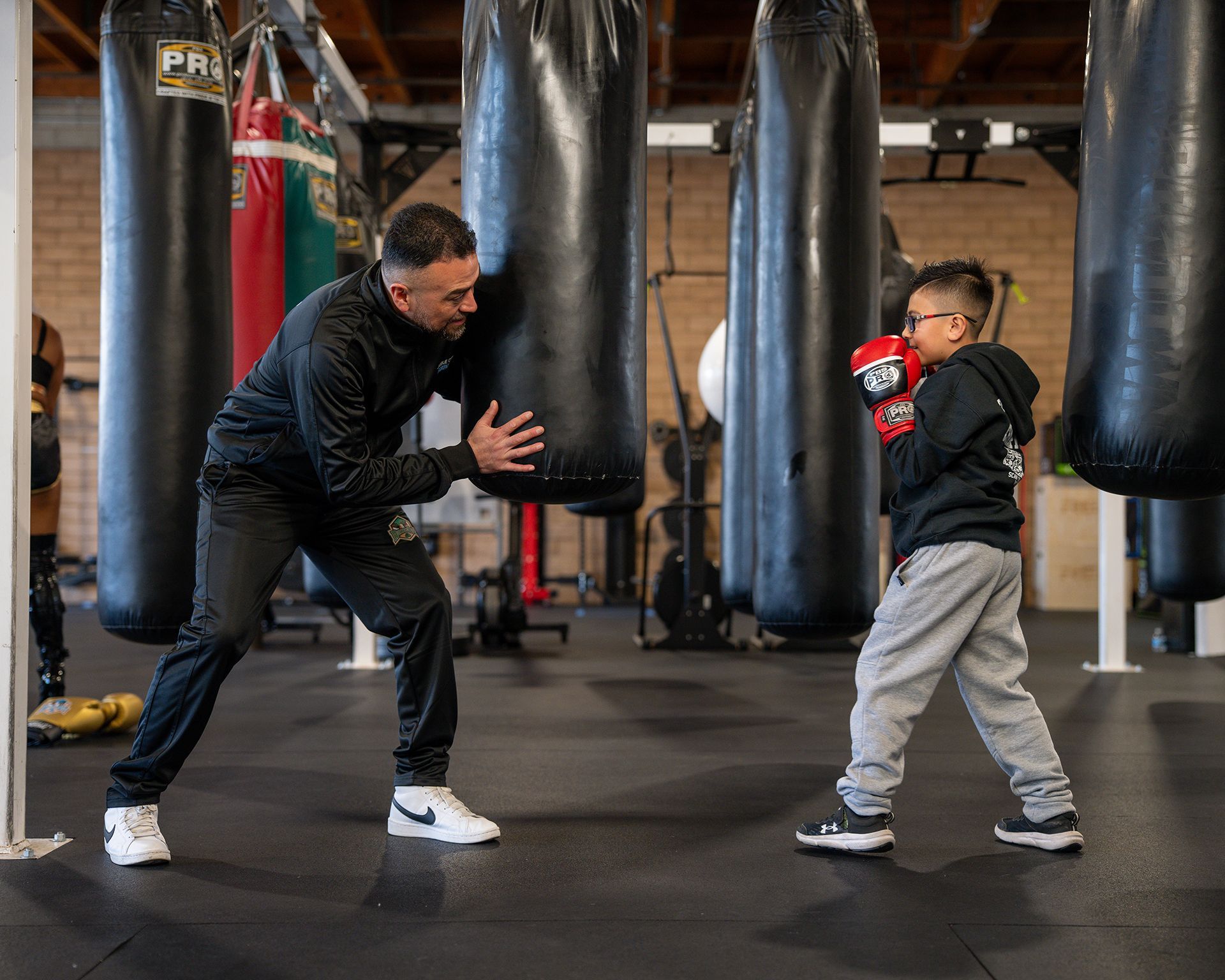 A man coaches a young boy wearing boxing gloves, at a gym with heavy bags, preparing to practice boxing.