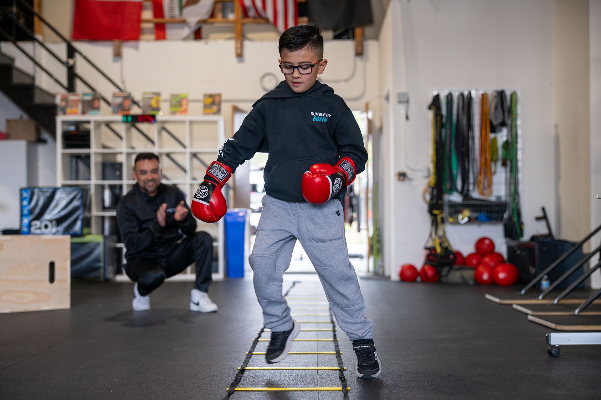 Boy with boxing gloves exercises on agility ladder; coach watches. Gym setting.