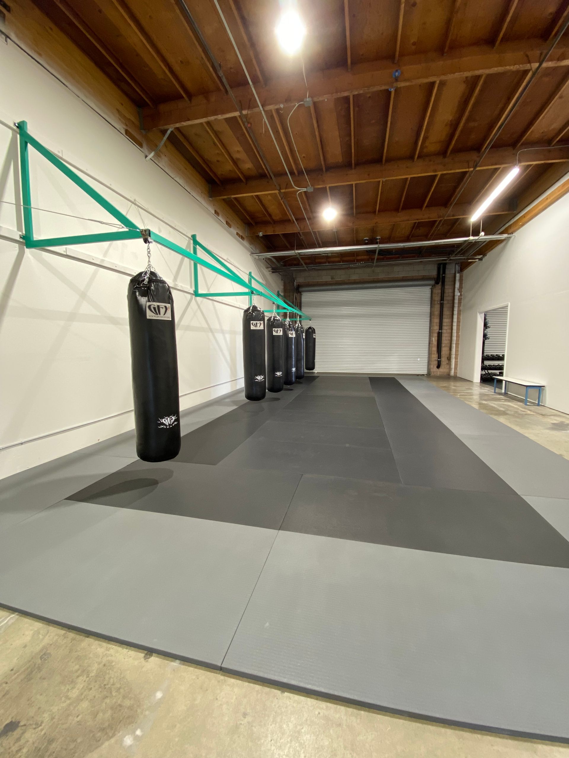 Boxing gym with black punching bags, gray mats, and a roll-up door.