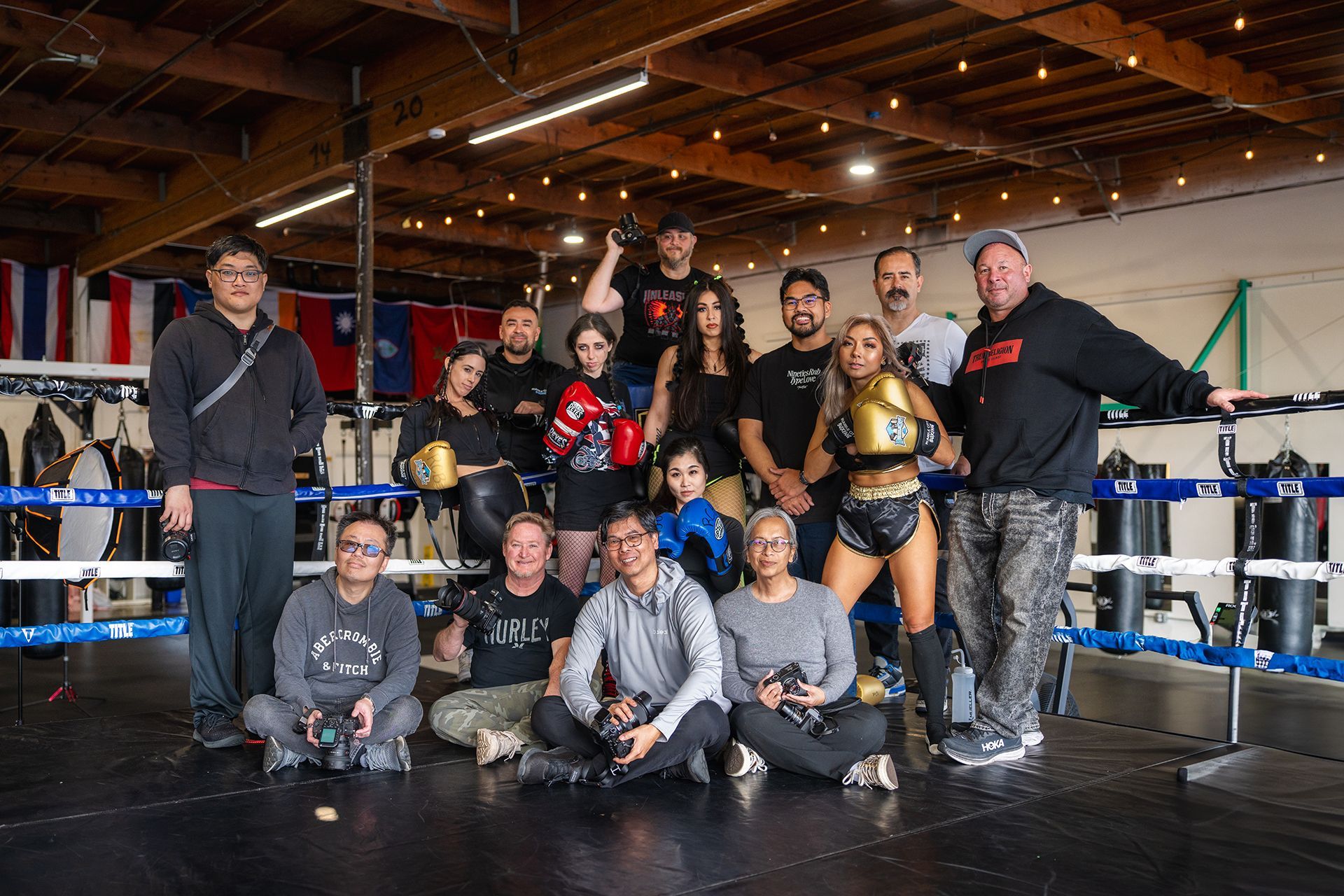 Group posing in boxing ring; diverse people, some wearing boxing gear, smiles. Flags in background, gym setting.