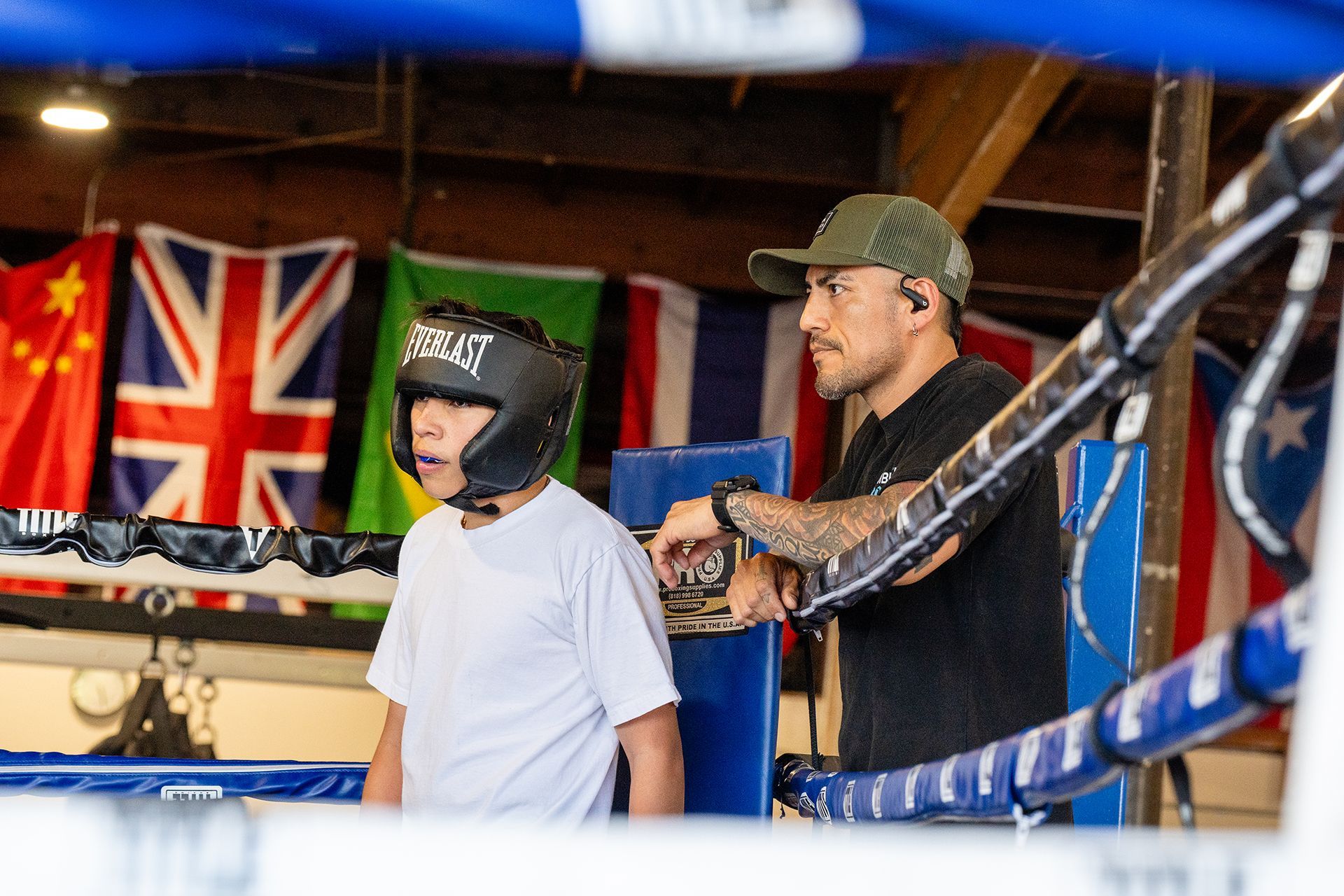 Boxer in protective headgear listens to a coach inside boxing ring. Flags hang in the background.